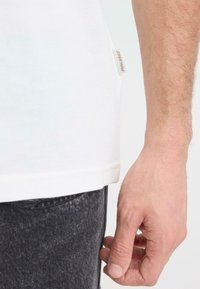 T-shirt blanc en coton avec tissu texturé, comportant une petite étiquette sur le côté gauche. Une main saisit l'ourlet, contrastant avec un jean en denim foncé.