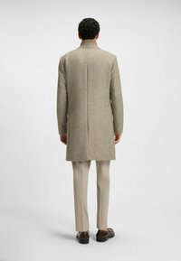 Beige checkered overcoat with a high collar, straight cut, and tailored fit, paired with light-colored trousers and brown shoes.