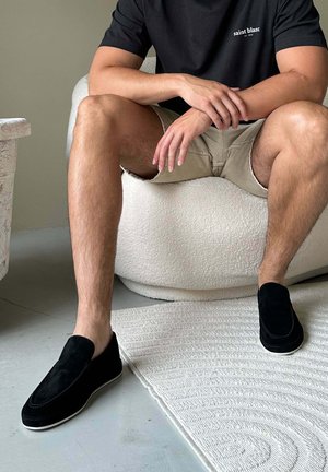 Man draagt zwarte instapschoenen, beige korte broek en zwart T-shirt, zittend op een witte, getextureerde fauteuil met handen naast elkaar rustend.