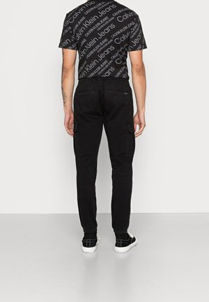 Man wearing black Calvin Klein Jeans t-shirt and black cargo pants, standing facing away on gray floor with white background.