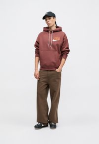 Hnedá mikina s kapucňou a predným vreckom, s bielymi šnúrkami, s oranžovým logom. Hnedé nohavice s širokými nohavicami a čierne tenisky dokončujú tento outfit.