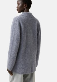 Woman wearing a soft, long-sleeve, light gray fuzzy sweater and light gray pants, viewed from the back and side profile.