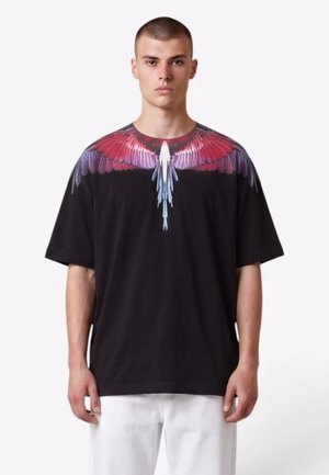 Giovane uomo che indossa una t-shirt nera con un disegno colorato di ali di uccello che si estende sulle spalle e sul petto, abbinata a pantaloni bianchi.