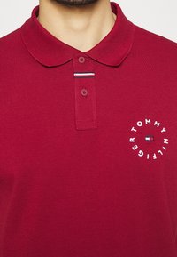 Bordowa koszulka polo z teksturowanym materiałem, kołnierzykiem, dwoma guzikami oraz białym nadrukowanym logo z kolorowym akcentem.