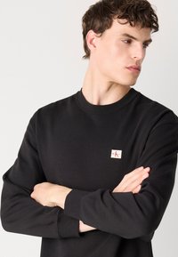 Zwarte sweatshirt met lange mouwen, gemaakt van zachte stof, met een ronde halslijn en een klein logo-opdracht op de borst. Eenvoudig design met een comfortabele pasvorm.