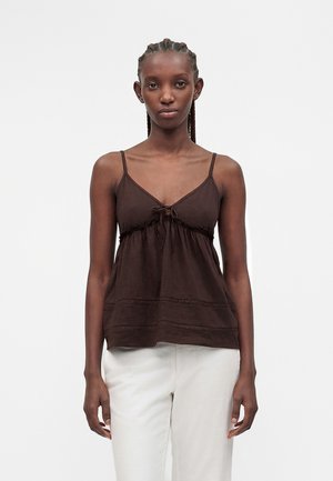 Femme aux cheveux tressés porte un haut sans manches marron foncé avec de fines bretelles et un pantalon blanc sur un fond blanc uni.