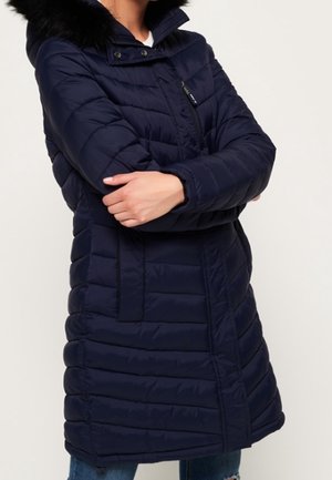 Kvinde iført en mørkeblå quiltet lang parka med sort pelsbeklædt hætte, lynlås og trykknapper, stående med armene korsede.