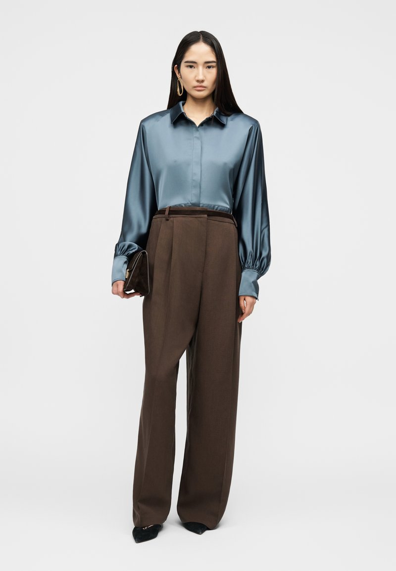 Femme portant une blouse en satin bleue à manches ballon, un pantalon large marron taille haute, tenant une pochette marron, debout.