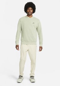 Grön sweatshirt med rund halsringning, texturerat tyg och en svart Nike-logotyp. Matchas med ljusbeiga byxor och atletiska skor.