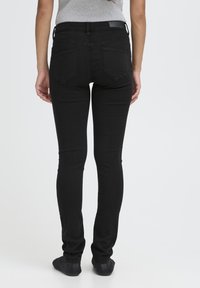 Persona de pie descalza vistiendo jeans ajustados negros y una blusa sin mangas gris, vista desde atrás contra un fondo blanco liso.