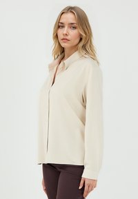 Beige Langarmhemd mit Button-Down-Kragen, glatter Textur und subtiler Glanz. Figurbetontes Design mit Knöpfen über die Vorderseite.