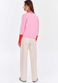 Różowo-czerwony sweter w blokach kolorów z długimi rękawami, prążkowanymi mankietami i okrągłym dekoltem, zestawiony z szerokimi spodniami w kolorze ecru. Miękka tekstura.
