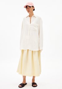 Hellbeige, oversized Hemdbluse mit einer Brusttasche, kombiniert mit einem plissierten gelben Midirock und schwarzen Slipper-Sandalen. Akzentuiert mit einer pinken Baseballmütze.