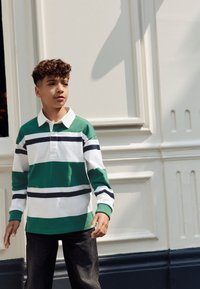 Rugbyové tričko s pruhy v zelené a bílé barvě s námořnicky modrými detaily, dlouhými rukávy a bílým límcem. Nošeno s tmavými džínovými kalhotami.