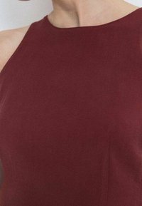 Robe sans manches bordeaux avec un col montant, une texture lisse, et une coupe ajustée soulignant une silhouette élancée.