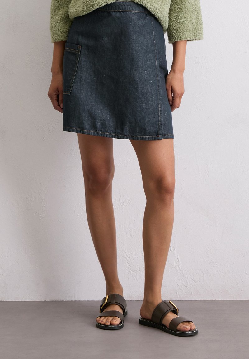 Jupe en denim bleu foncé avec une poche avant, associée à des sandales marron avec un accent doré et une semelle plate. Pull vert à texture douce.