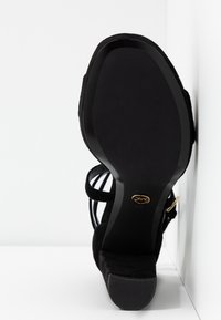 Sandale à talons hauts en daim noir avec talon ouvert, fines lanières et semelle noire lisse ornée d'un logo doré.