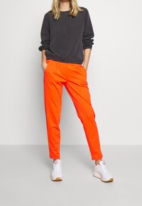 Felpa nera con polsini elasticizzati, abbinata a pantaloni jogger arancioni vivaci con tasche laterali e una gamba svasata. Sneaker bianche.