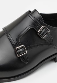 Schwarze Lederschuhe mit doppeltem Schnallenakzent, glatter Oberfläche und dezenten Nähdetails entlang des Obermaterials und der Sohle.