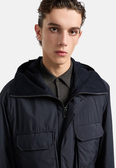 Giacca puffer blu navy con cappuccio foderato in pile, zip frontale e due tasche sul petto. Camicia grigia testurizzata sotto, con colletto a bottoni.