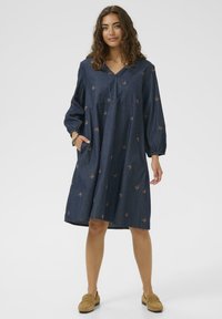 Frau in einem lockeren dunkelblauen Kleid mit Schmetterlingsmustern und braunen Mokassins, die mit einer Hand in der Tasche vor einem weißen Hintergrund steht.