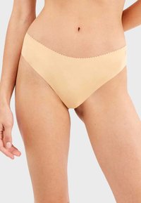 Bikini de corte bajo en color beige, sin costuras, con bordes ondulados, textura suave y cobertura mínima para mayor comodidad y discreción.