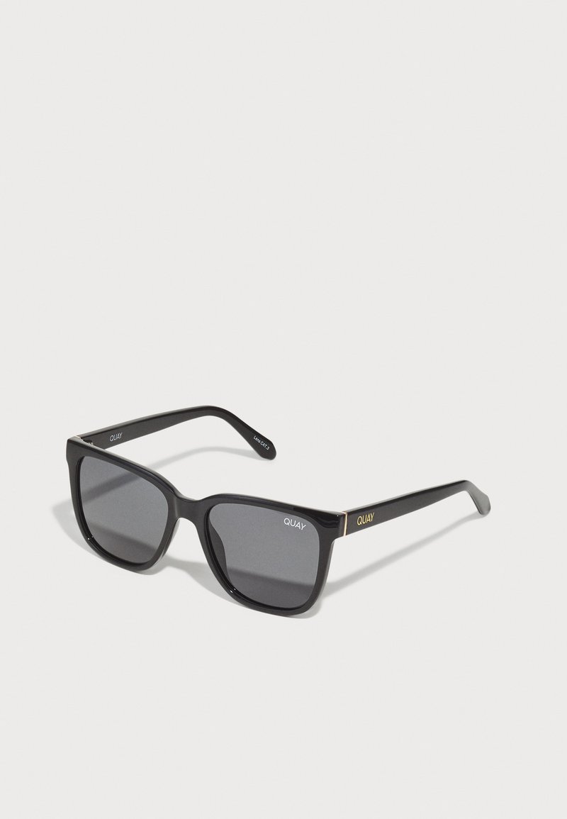 Gafas de sol cuadradas negras con lentes oscuros, acabado mate, acentos dorados en las patillas y logo grabado. Diseño angular, marco ancho.