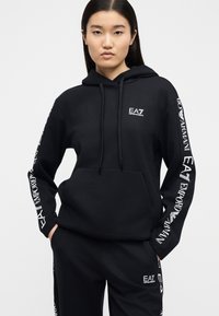 Νεαρή γυναίκα που φοράει μαύρο φούτερ Emporio Armani EA7 με κουκούλα και αντίστοιχη φόρμα με λογότυπα στα μανίκια και στο στήθος, στέκεται με τα χέρια στις τσέπες.