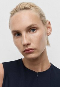 Detailní záběr na osobu s světlými blond vlasy, světlou pletí a pihami, která má na sobě námořnickou bezrukávovou blůzu s diskrétním zipem na výstřihu.