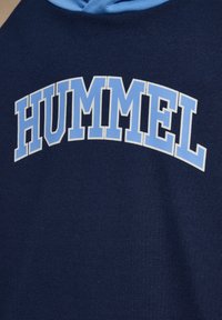 Granatowa bluza z kapturem z pogrubionym napisem "HUMMEL" w jasno niebieskim i białym kolorze, ułożonym łukiem na klatce piersiowej, z jasnoniebieską podszewką kaptura.