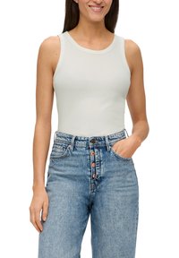 Donna con una canotta bianca senza maniche e jeans a vita alta blu con chiusura a bottoni, una mano in tasca, che sorride su uno sfondo bianco.