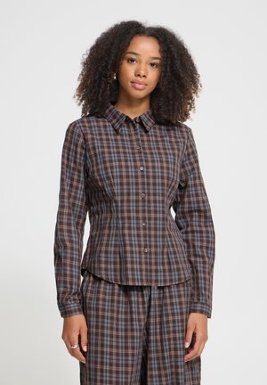 Femme aux cheveux bouclés portant une chemise à carreaux marron et bleue boutonnée et un pantalon assorti, debout devant un fond uni clair.