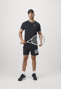 Černé sportovní tričko s bílým olemováním, černé kraťasy s nápisem "NIKE" v bílé barvě, drží tenisovou raketu, má na hlavě černou kšiltovku a černé tenisky.