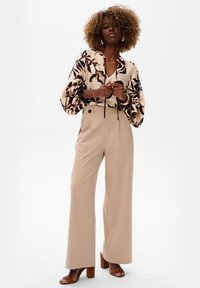 Beige high-waisted broeken gecombineerd met een crèmekleurige blouse met zwarte bloemenpatronen. De blouse heeft pofmouwen en een koord. Bruine blokhakken.
