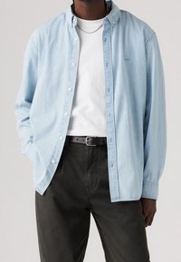 Man in een lichtblauw overhemd over een wit T-shirt, met een zwarte riem en donkere broek, met zijn handen in zijn zakken op een neutrale achtergrond.