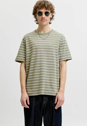 Ung mand med runde solbriller, en lysegrøn og hvid stribet t-shirt, mørke bukser og en tynd kædehalskæde mod en ensfarvet baggrund.