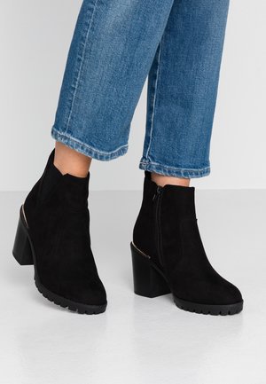 Bottines en daim noir à talons épais portées avec un jean bleu court sur un sol clair et un fond uni.