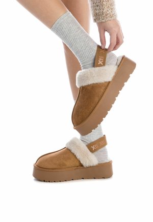 Zapatos marrones de ante tipo slip-on con una banda de frenoso crema esponjoso, suela gruesa texturizada y correa color tostado que presenta el logo "Xti" en marrón claro.