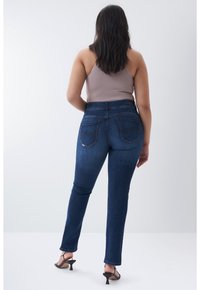 Mujer con cabello largo y oscuro, vestida con una blusa sin mangas color topo, jeans azul oscuro y sandalias negras de tacón, de pie de espaldas sobre un fondo blanco.