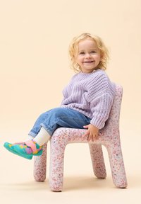 Niño sentado en una silla con manchas rosas hecha de un material texturado, vistiendo un suéter de punto lila, jeans azules y zapatos coloridos.