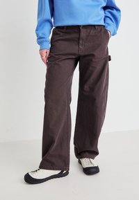 Brune vide bukser laget av mykt stoff, med knappelukking og en sidelomme. Kombinert med lyse sneakers.