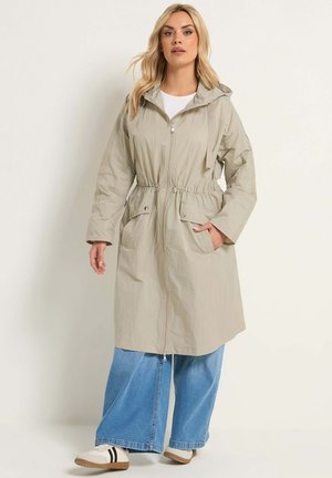 Donna che indossa un lungo giaccone beige con cappuccio e tasche frontali, jeans larghi blu e scarpe da ginnastica bianche con strisce nere, in piedi.