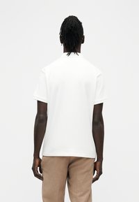 Homme à la peau foncée et dreadlocks, portant un t-shirt blanc à manches courtes côtelé et un pantalon beige, debout de dos sur un fond uni.