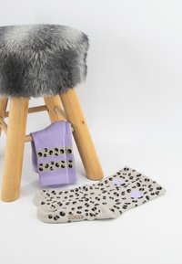 Deux paires de chaussettes à motifs : l'une violet clair avec des taches de léopard et l'autre crème avec des taches noires et violettes, placées près d'un tabouret en bois.