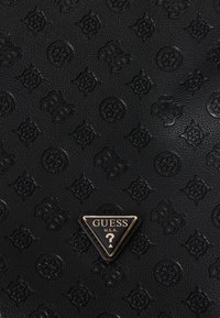 Superfície de couro texturizado preto com padrões florais em relevo e "GS" entrelaçado, apresentando uma placa de logotipo triangular da Guess U.S.A. com borda dourada.