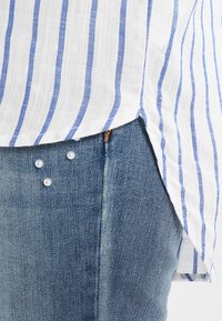 Camicia a righe bianca e blu con orlo arrotondato, abbinata a jeans in denim blu decorati con tre perline a forma di perla.
