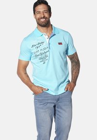 Hellblaues Poloshirt mit weicher Textur, ausgestattet mit einer Drei-Knopf-Leiste, Logo-Patch und gedruckten Grafiken auf der Vorderseite. Getragen mit hellen Jeans.