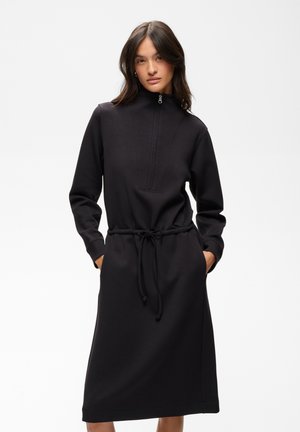 Femme portant une longue robe noire avec une fermeture éclair à l'avant, un col montant et une taille avec cordon, mains dans les poches sur fond blanc uni.