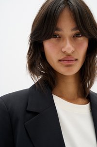 Black blazer over a white top, featuring a structured collar. Hair is dark and tousled, with a smooth skin texture and neutral tones.