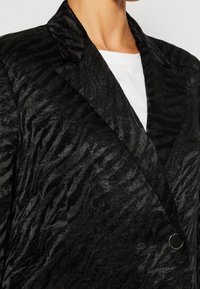 Blazer nero con texture e motivo a onde, rever a punta e chiusura con un singolo bottone rotondo, abbinato a una maglietta bianca.